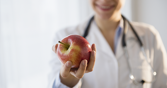 A smiling doctor wearing a white coat and stethoscope offers her patient a fresh apple while they talk comprehensive care