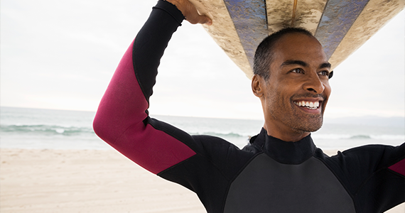 A smiling man is using his paid time off to catch some waves. He is wearing a wetsuit and holds his surfboard over his head