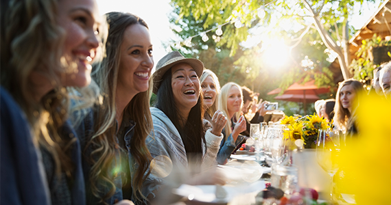 Friends and family sit and converse at a long table as they prepare to share a meal in celebration of a summer holiday